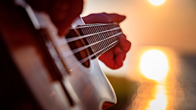 A close-up of a guitar being played, strings and fretboard in focus, with a warm sunset glow reflecting off the water in the background.