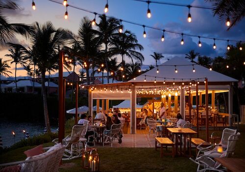 A tropical outdoor dining area at dusk with string lights, palm trees, and warmly lit tables under a canopy; guests enjoy a cozy evening by the water.