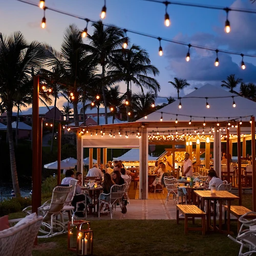 A tropical outdoor dining area at dusk with string lights, palm trees, and warmly lit tables under a canopy; guests enjoy a cozy evening by the water.