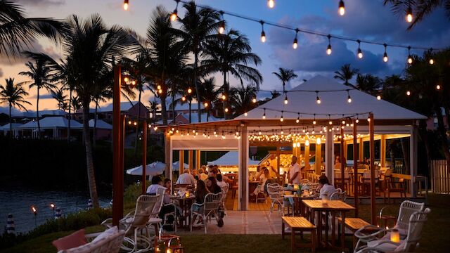 A tropical outdoor dining area at dusk with string lights, palm trees, and warmly lit tables under a canopy; guests enjoy a cozy evening by the water.