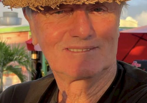A smiling man in a straw hat and black shirt is sitting outdoors with trees and a building in the background, looking content.