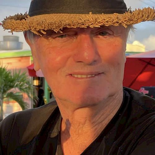 A smiling man in a straw hat and black shirt is sitting outdoors with trees and a building in the background, looking content.