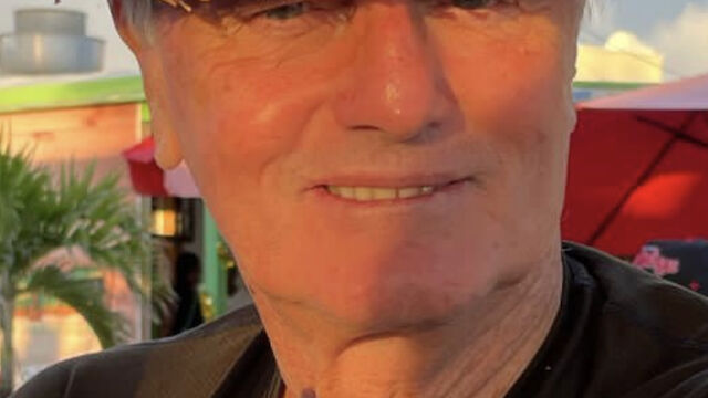 A smiling man in a straw hat and black shirt is sitting outdoors with trees and a building in the background, looking content.