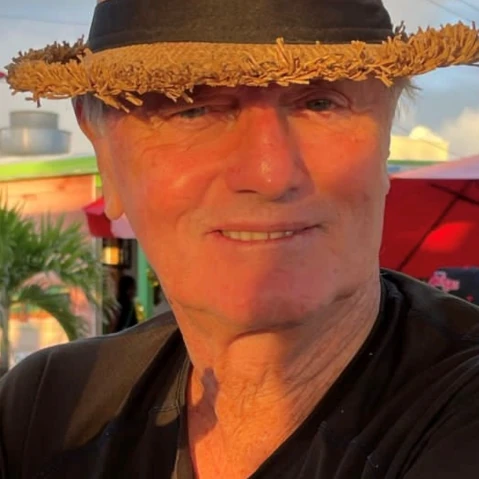 A man wearing a straw hat with a black band smiles for a sunny outdoor selfie.