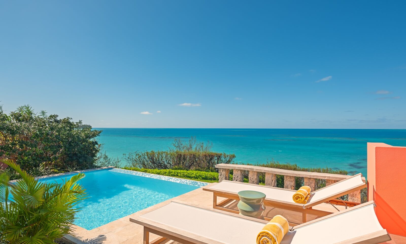 A sunny oceanfront pool area with clear blue water, lounge chairs, rolled towels, and a relaxing seaside backdrop.