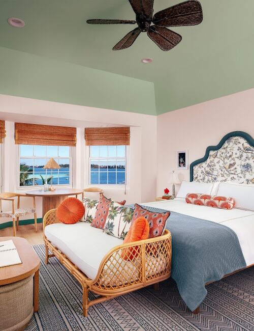 A bright coastal bedroom with a pastel green ceiling, mint walls, a woven headboard bed, wicker seating, and large windows overlooking the sea, cozy and airy.