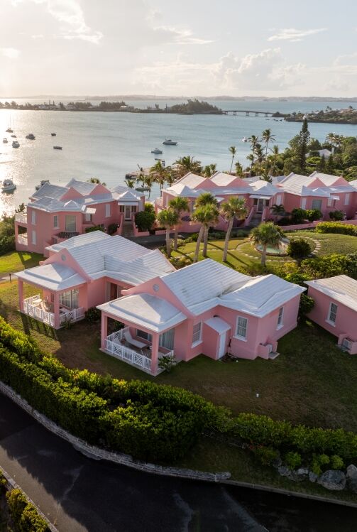 Aerial view of a pink resort by a calm bay, palm trees, pastel villas, and a winding road, with sparkling water and distant horizon.