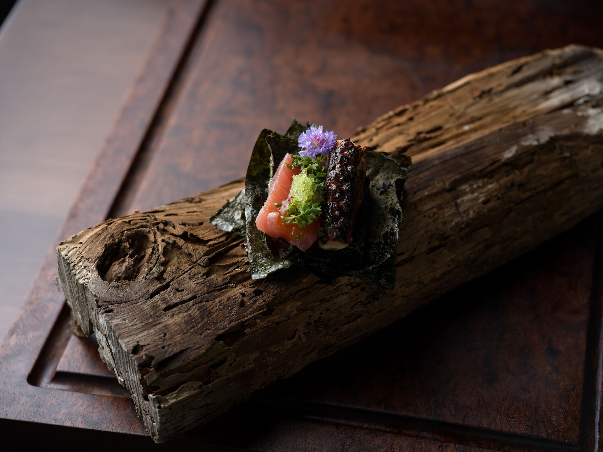 A gourmet plate of seared meat and pale seafood, topped with greens and a purple flower, served on a wooden slab.