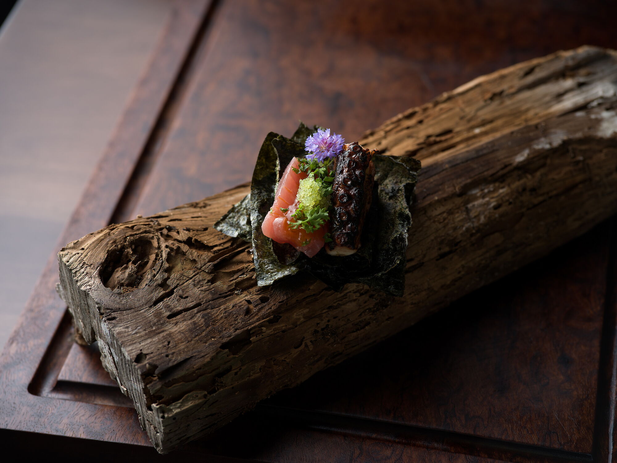 A gourmet plate of seared meat and pale seafood, topped with greens and a purple flower, served on a wooden slab.