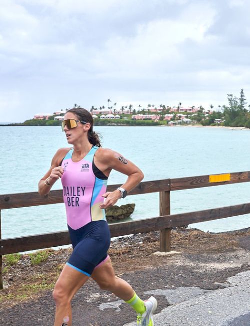 A person is running along a scenic coastal path, with the ocean on one side and trees in the distance, near a yellow cone on the road.
