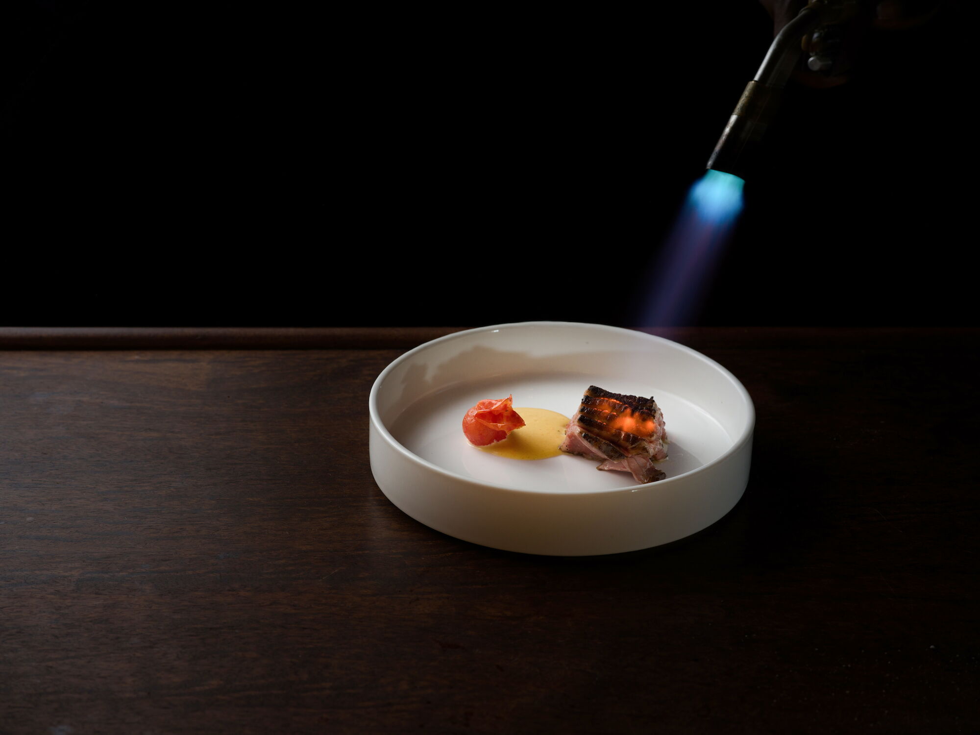 A lit gas torch hovers over a plated gourmet dish with bacon-wrapped meat and a small garnish on a white bowl.
