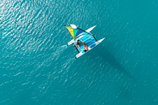 A top-down view of two people sailing on a trimaran in clear blue water, with a colorful sail and bright sunlight casting shadows on the water's surface.