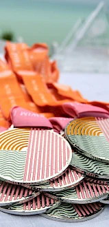 A table with a stack of colorful medals with orange and pink lanyards, arranged neatly against a backdrop of a blurry outdoor setting.