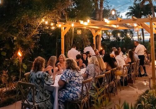 An outdoor evening dinner party with guests seated at tables under warm string lights and tiki-style decor.