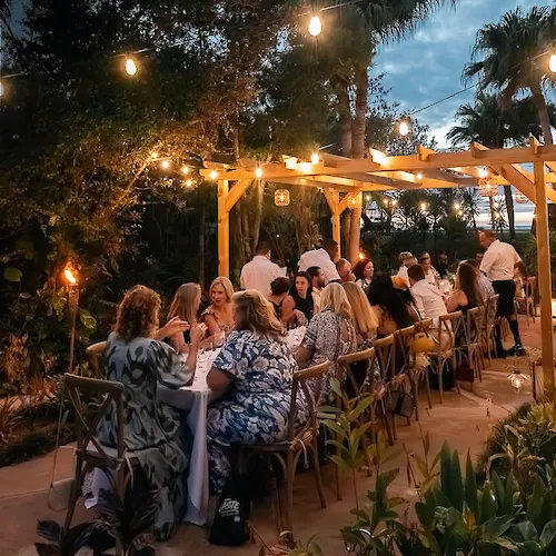An outdoor evening dinner party with guests seated at tables under warm string lights and tiki-style decor.