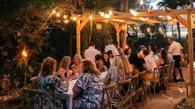 An outdoor evening dinner party with guests seated at tables under warm string lights and tiki-style decor.