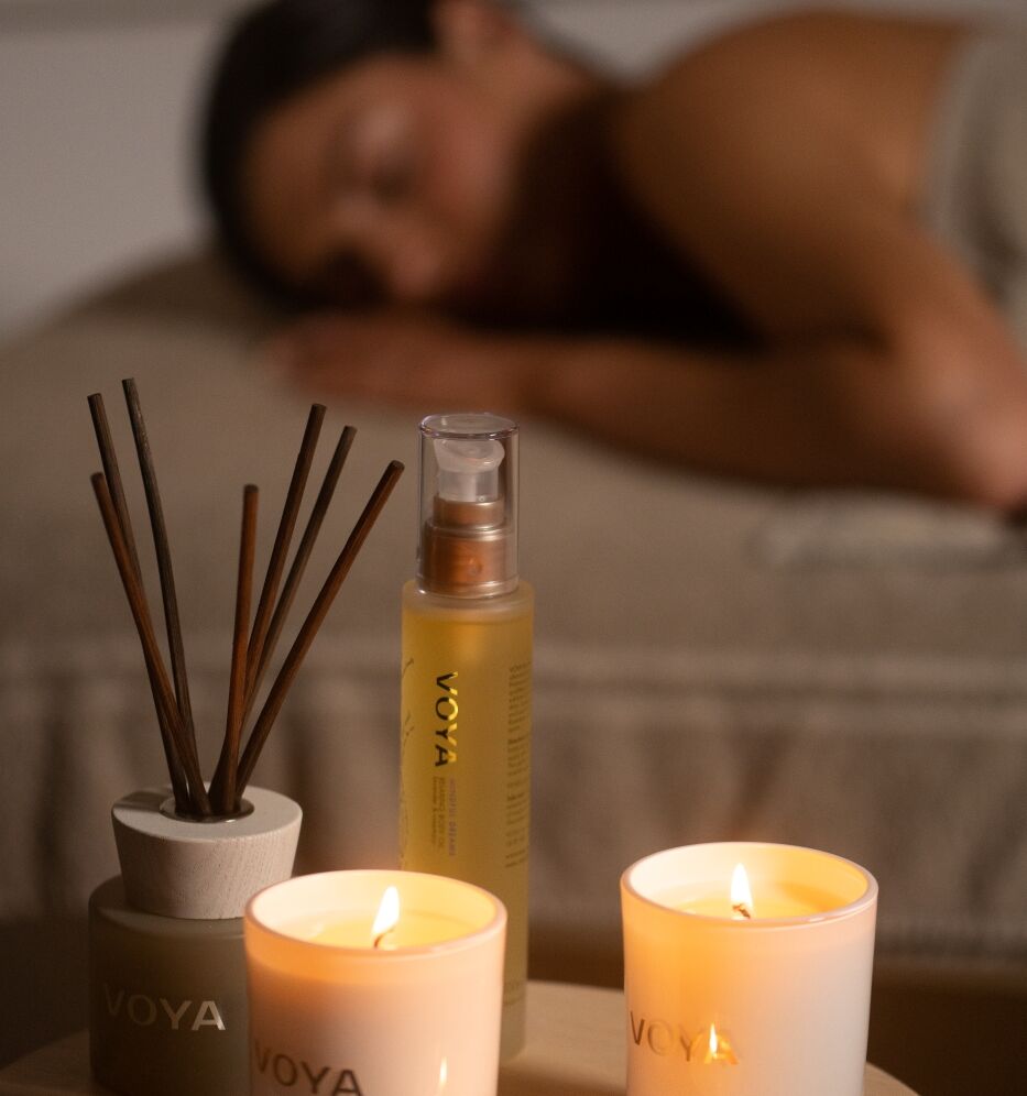 Lit VOYA candles and a reed diffuser on a wooden tray, with a blurred person resting in the background, glowing warmly.