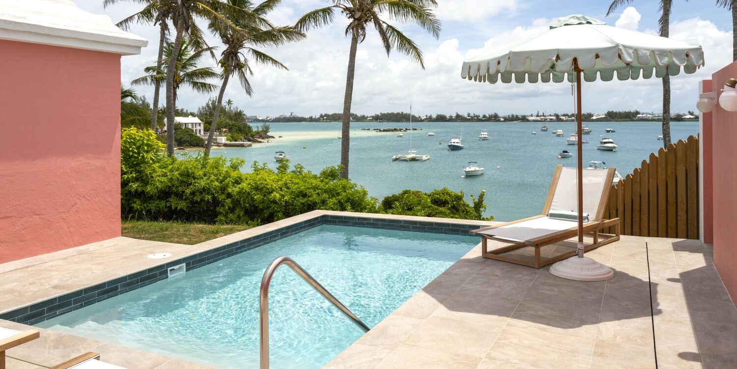 A sunny resort scene with a small pool, palm trees, a waterfront view, boats on the water, and a white umbrella by a lounge chair.
