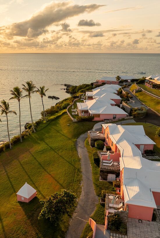 Sunset over a tropical resort: palm-lined lawn, pastel cottages along the coast, calm sea, golden sky, peaceful vacation vibes.