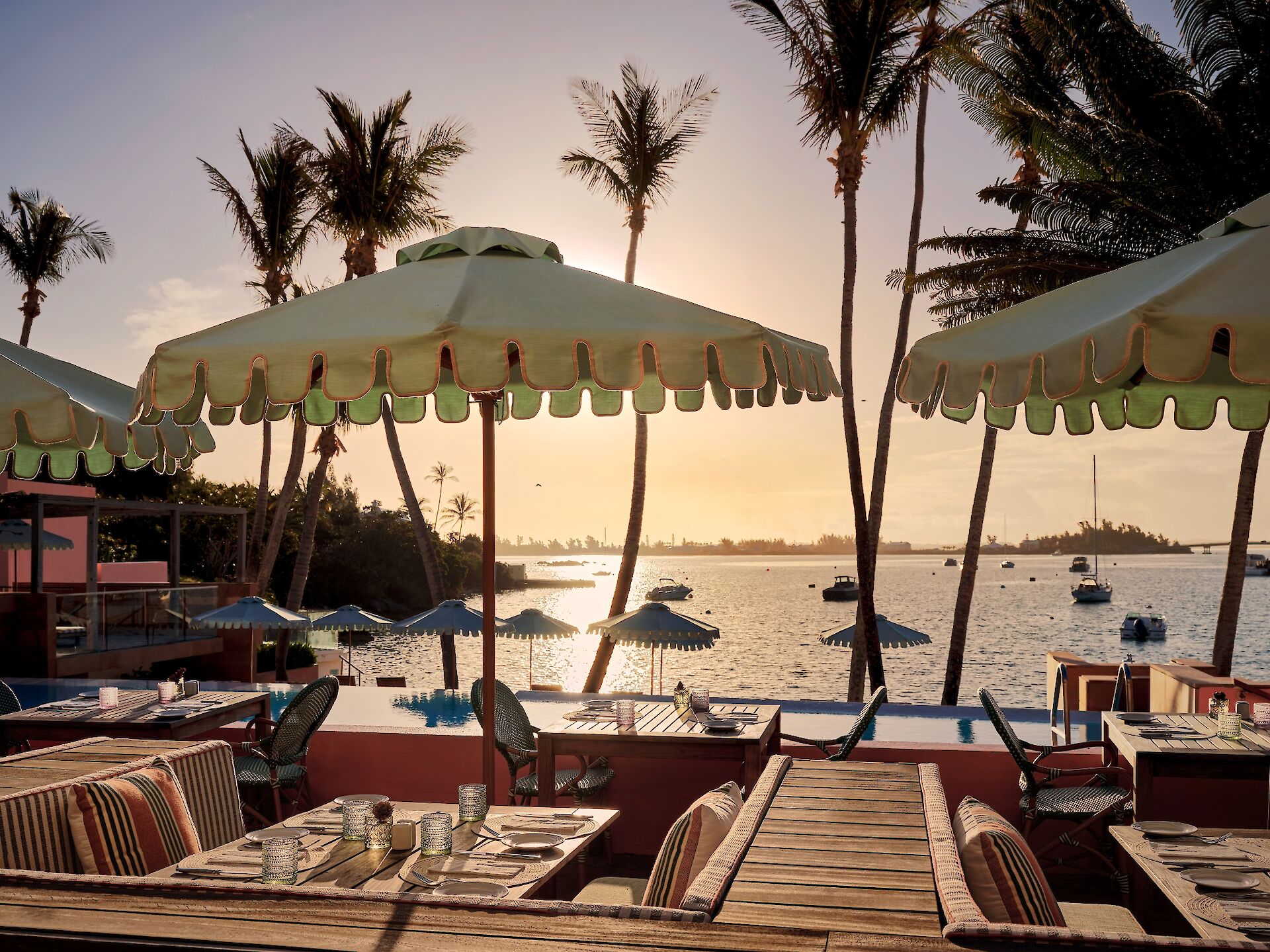 A tropical seaside restaurant view with palm trees, striped chairs, and colorful cabana umbrellas over calm water at sunset.