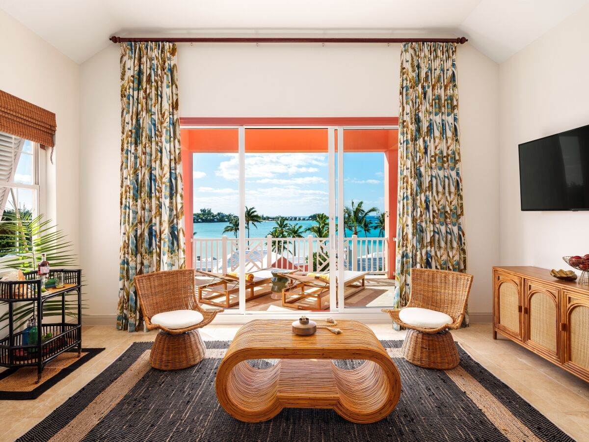 Sunlit tropical living room with wicker chairs and coffee table, open balcony doors showing palms and ocean under blue sky.