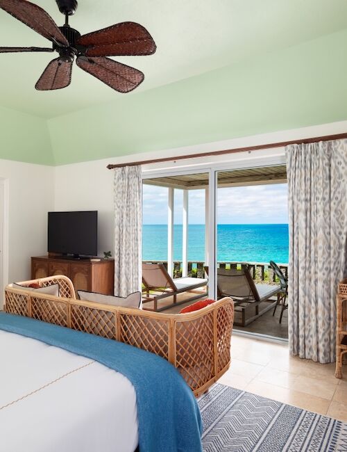 A bright seaside bedroom with a ceiling fan, rattan seating, and large sliding doors opening to a blue ocean view.
