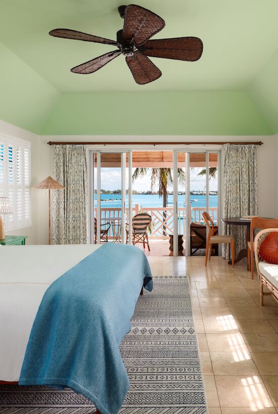 A cozy bedroom with a large bed, ceiling fan, and tropical decor, overlooking a balcony with an ocean view through glass doors.
