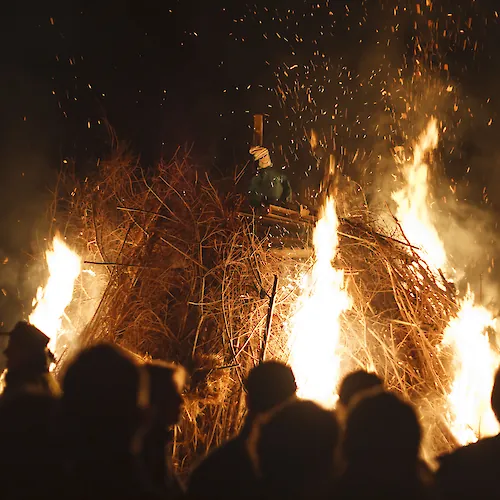 Large nighttime bonfire surrounded by a crowd, with bright flames and sparks rising into the dark sky.