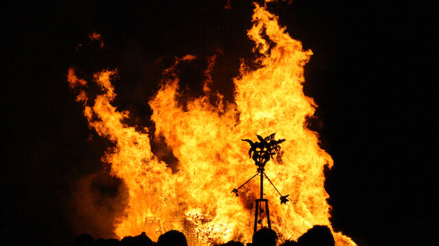 A large bonfire prominently burns in the night, with a silhouetted metal structure and crowd of shadows visible in the foreground.