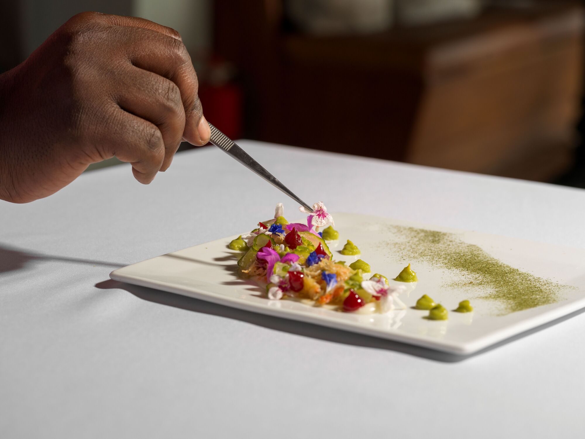 A person is elegantly arranging colorful food on a rectangular white plate with tweezers, showcasing haute cuisine or fine dining art.