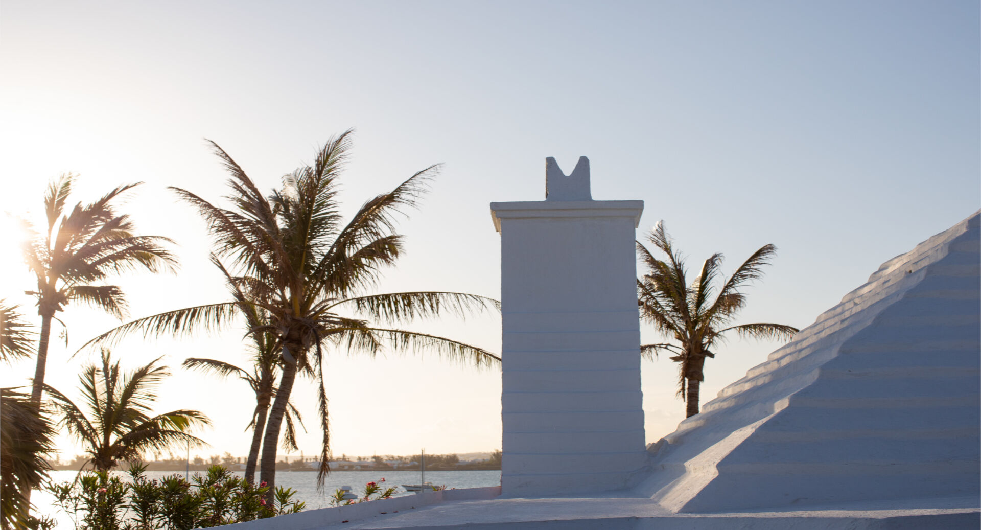 A white pillar tower with a horseshoe top stands beside palm trees and a sandy walkway at sunset by the sea.