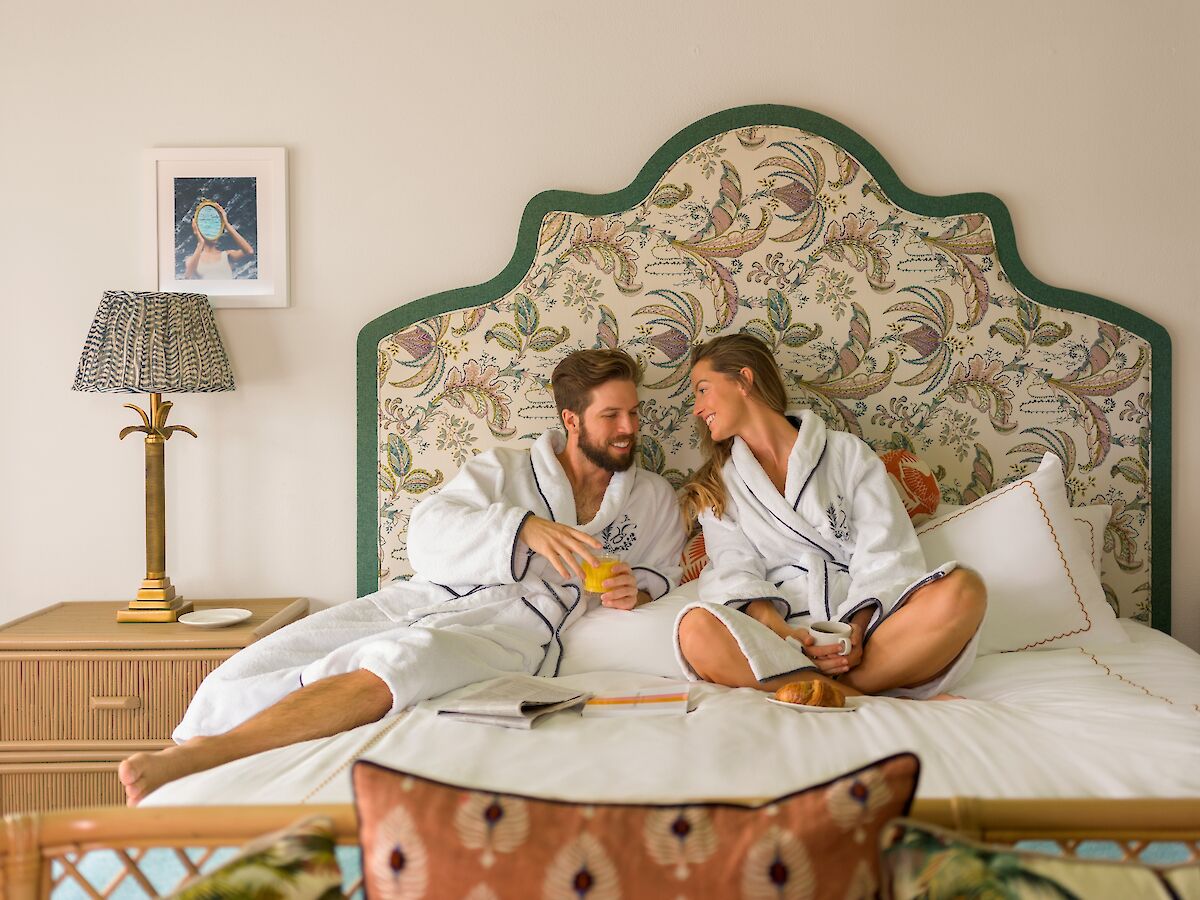 A couple in white robes sit on a bed, smiling and chatting, with a floral headboard, nightstand lamp, and a small framed photo on the wall.