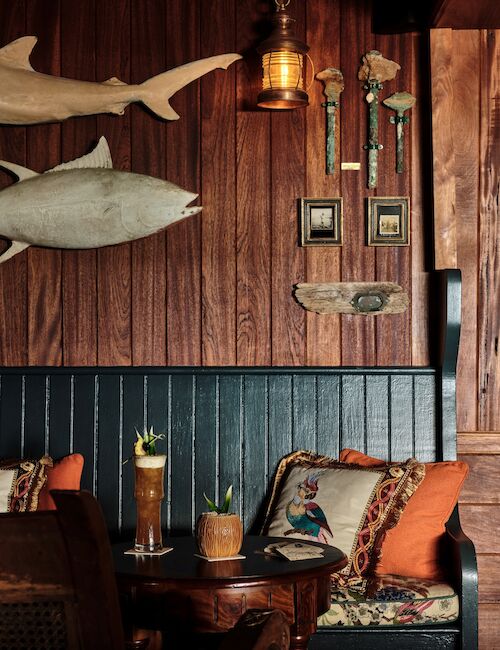 Cozy booth-style caf&eacute; with dark wood walls, a teal bench, orange pillows, a round table, potted plants, wall-mounted fish and rustic decor, warm lighting.