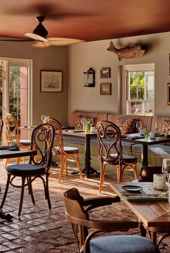 A cozy, rustic dining room with a view of trees outside; mismatched wooden tables and chairs, a brick fireplace, framed art, and soft natural light.