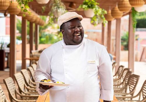A smiling chef in a white jacket and hat holds a plate of food in an outdoor cafe with green plants overhead.