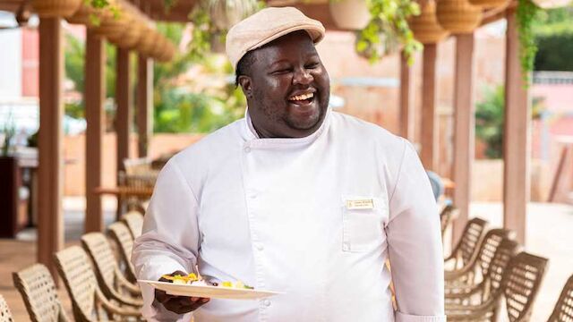 A smiling chef in a white jacket and hat holds a plate of food in an outdoor cafe with green plants overhead.