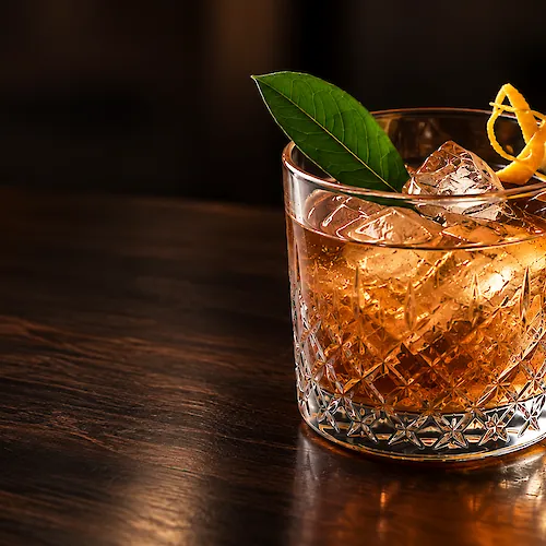 A crystal tumbler of amber cocktail with ice, garnished with a green leaf and curled orange peel.