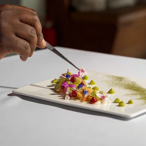 A hand uses tweezers to arrange a colorful, artistic dish on a white rectangular plate, showcasing a fine dining presentation.