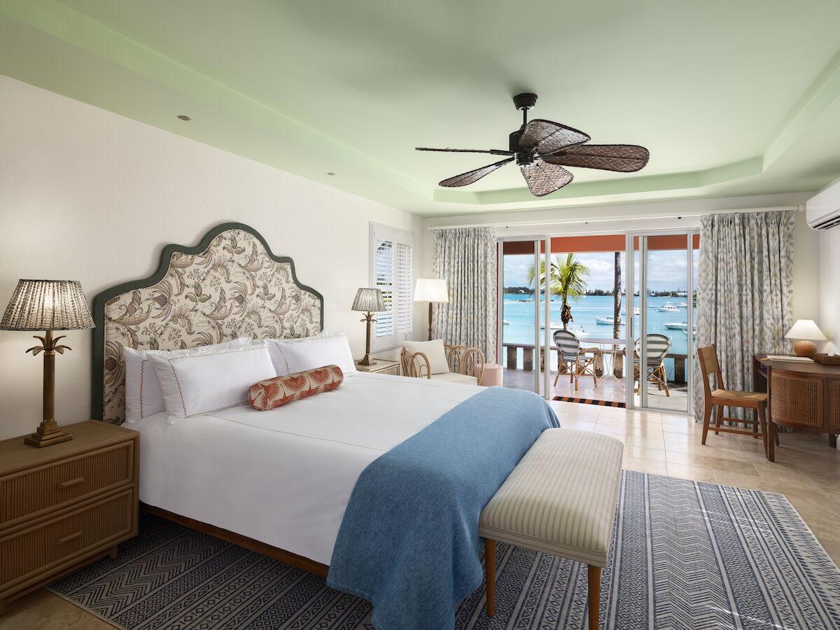 A cozy bedroom with a sea view features a bed, ceiling fan, lamps, and a desk. The balcony offers seating with ocean scenery beyond the glass doors.