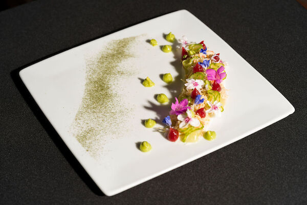 A gourmet dish on a square plate with colorful garnishes and green sauce dots, artfully arranged.