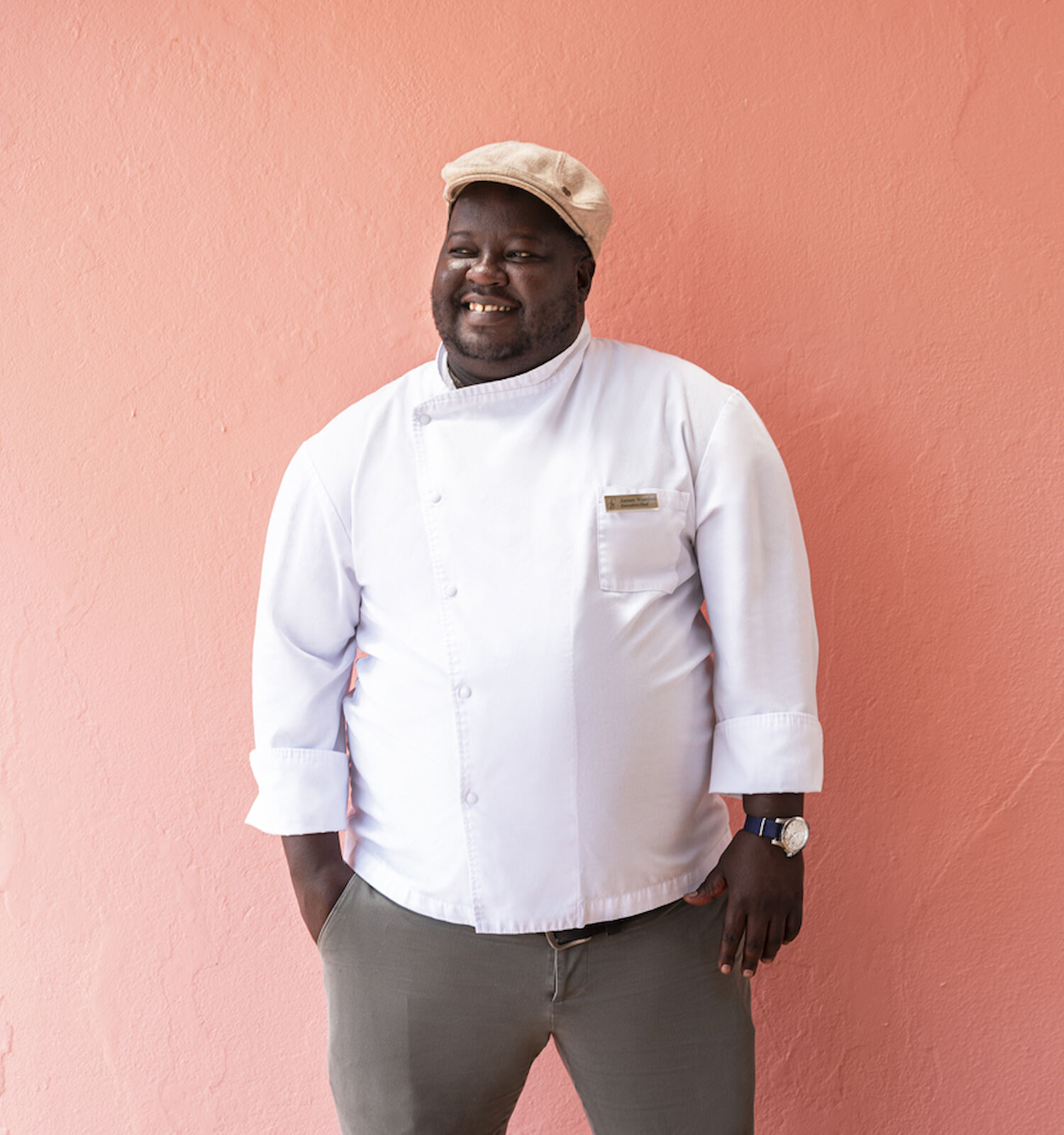 A person in a chef's uniform and cap is standing against a pink wall, smiling with hands in pockets.