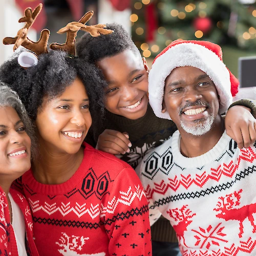 A family wearing festive sweaters and accessories takes a selfie in front of a decorated Christmas tree, smiling and celebrating the holiday.
