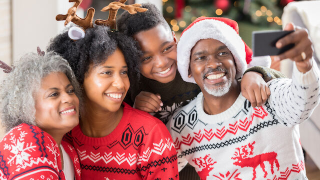 A family wearing festive sweaters and accessories takes a selfie in front of a decorated Christmas tree, smiling and celebrating the holiday.
