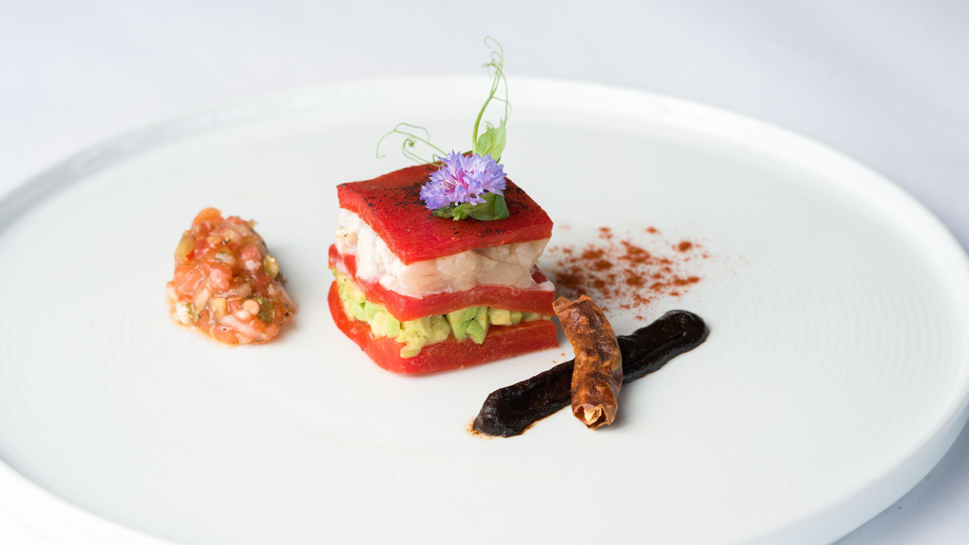 A plated gourmet entr&eacute;e with stacked red/white fish or seafood, avocado-like filling, garnishes, and an edible purple flower on a white plate.
