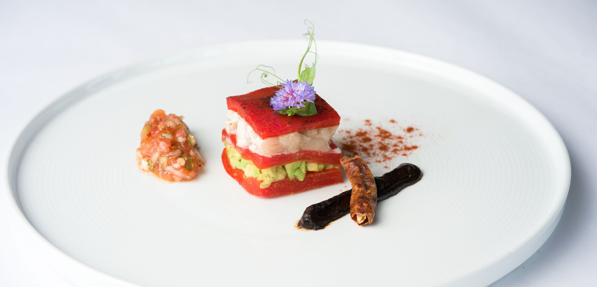 A plated gourmet entr&eacute;e with stacked red/white fish or seafood, avocado-like filling, garnishes, and an edible purple flower on a white plate.