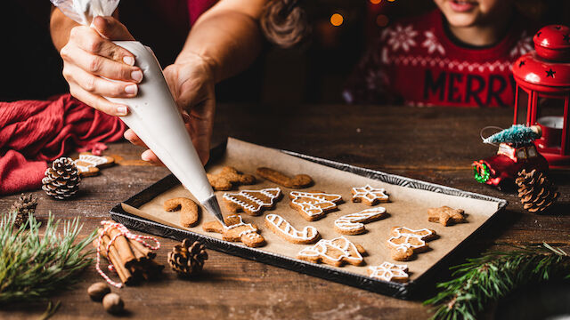A person is decorating Christmas-themed cookies with icing on a tray, surrounded by holiday decorations like pine cones and greenery.