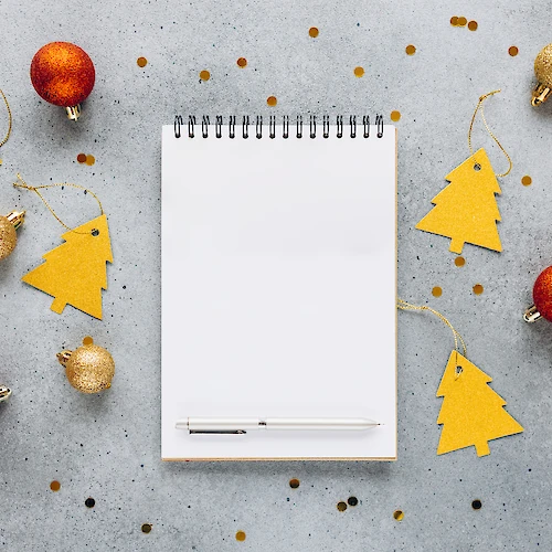 A spiral notebook and pen are surrounded by gold and red Christmas ornaments and tree-shaped tags on a gray surface, with scattered confetti.