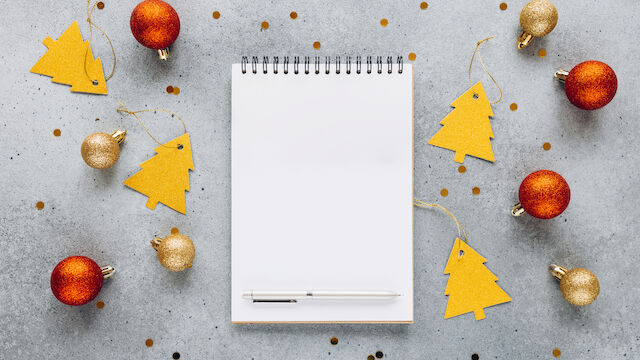 A spiral notebook and pen are surrounded by gold and red Christmas ornaments and tree-shaped tags on a gray surface, with scattered confetti.