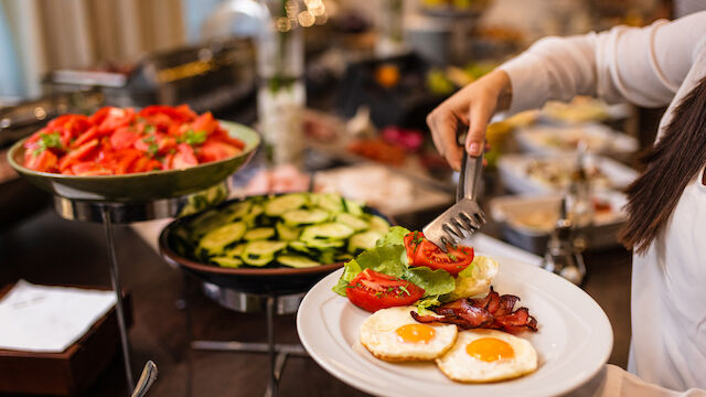 A person serves food onto a plate at a buffet, adding tomatoes and greens beside sunny-side-up eggs and bacon.