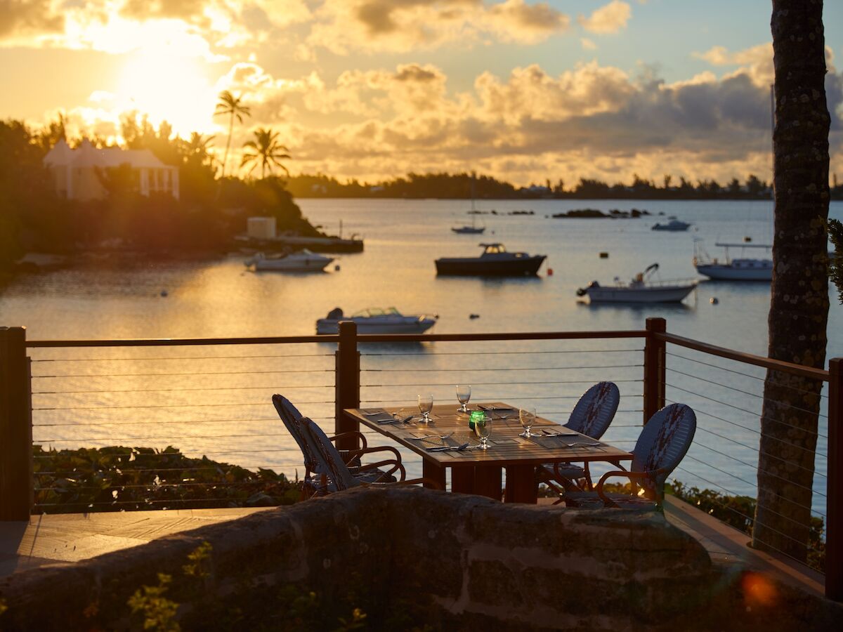 A relaxing seaside patio at sunset with boats gently floating on calm water and a cozy table for two overlooking the harbor.